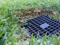 Czarna kratka odprowadzająca wodę z rynien na trawnik, otoczona zieloną trawą.