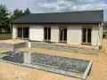 Dom w stanie surowym zamkniętym, z dachem i oknami. Wokół fundamenty i piasek.
