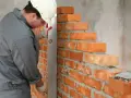 Murarz w kasku i rękawicach sprawdza pion muru z cegły, dbając o prawidłowe wymiary cegły i równość ścian.
