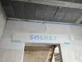 Ściana z bloczków z napisem "SOLBET" i pianką montażową. W tle widać sufit z pustaków i drabinę. Dykta nie jest widoczna.