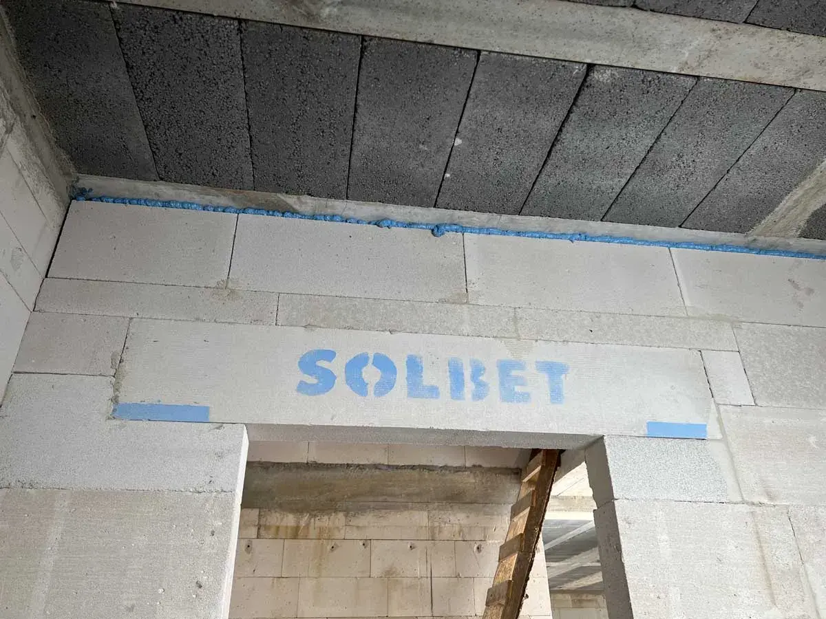 Ściana z bloczków z napisem "SOLBET" i pianką montażową. W tle widać sufit z pustaków i drabinę. Dykta nie jest widoczna.