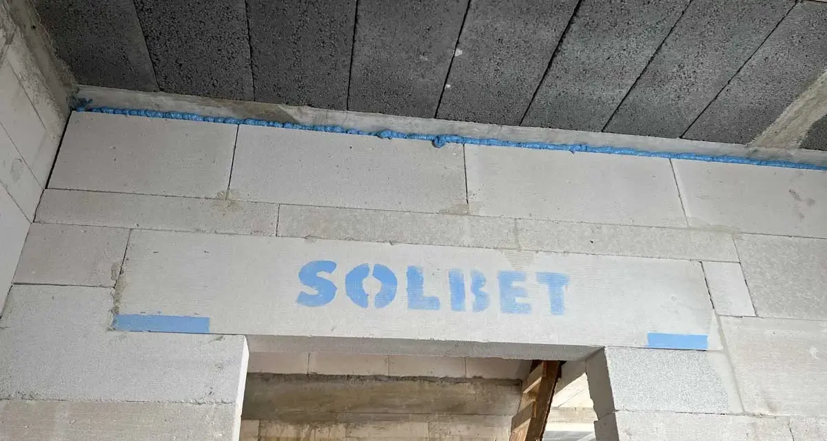 Ściana z bloczków z napisem "SOLBET" i pianką montażową. W tle widać sufit z pustaków i drabinę. Dykta nie jest widoczna.
