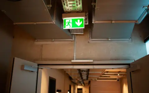Zielone światła awaryjne wskazują drogę ewakuacji w korytarzu z widocznymi instalacjami wentylacyjnymi i betonowymi ścianami.