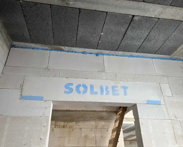 Ściana z bloczków z napisem "SOLBET" i pianką montażową. W tle widać sufit z pustaków i drabinę. Dykta nie jest widoczna.