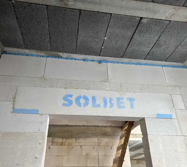 Ściana z bloczków z napisem "SOLBET" i pianką montażową. W tle widać sufit z pustaków i drabinę. Dykta nie jest widoczna.