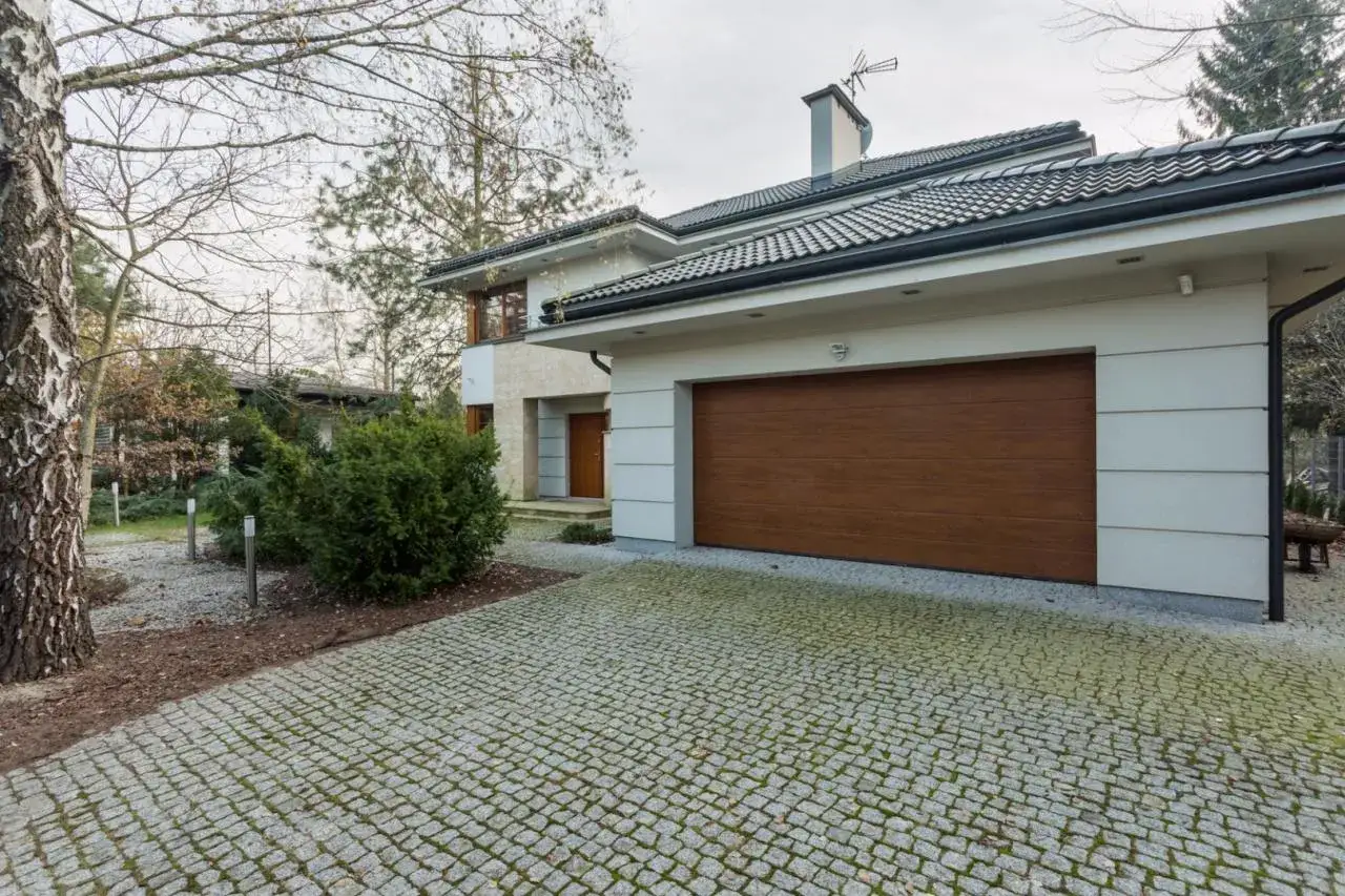 Nowoczesny dom z brązową bramą garażową i kamienną podbitką. Eleganckie boniowanie podkreśla fasadę, a podjazd z kostki brukowej dodaje uroku.