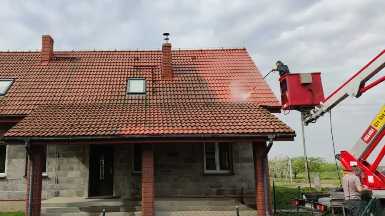 Pracownik z podnośnika myje dach domu. Woda pod ciśnieniem usuwa mech i brud z dachówki, przywracając jej dawny blask. Okapnik na dachu jest czysty i suchy.