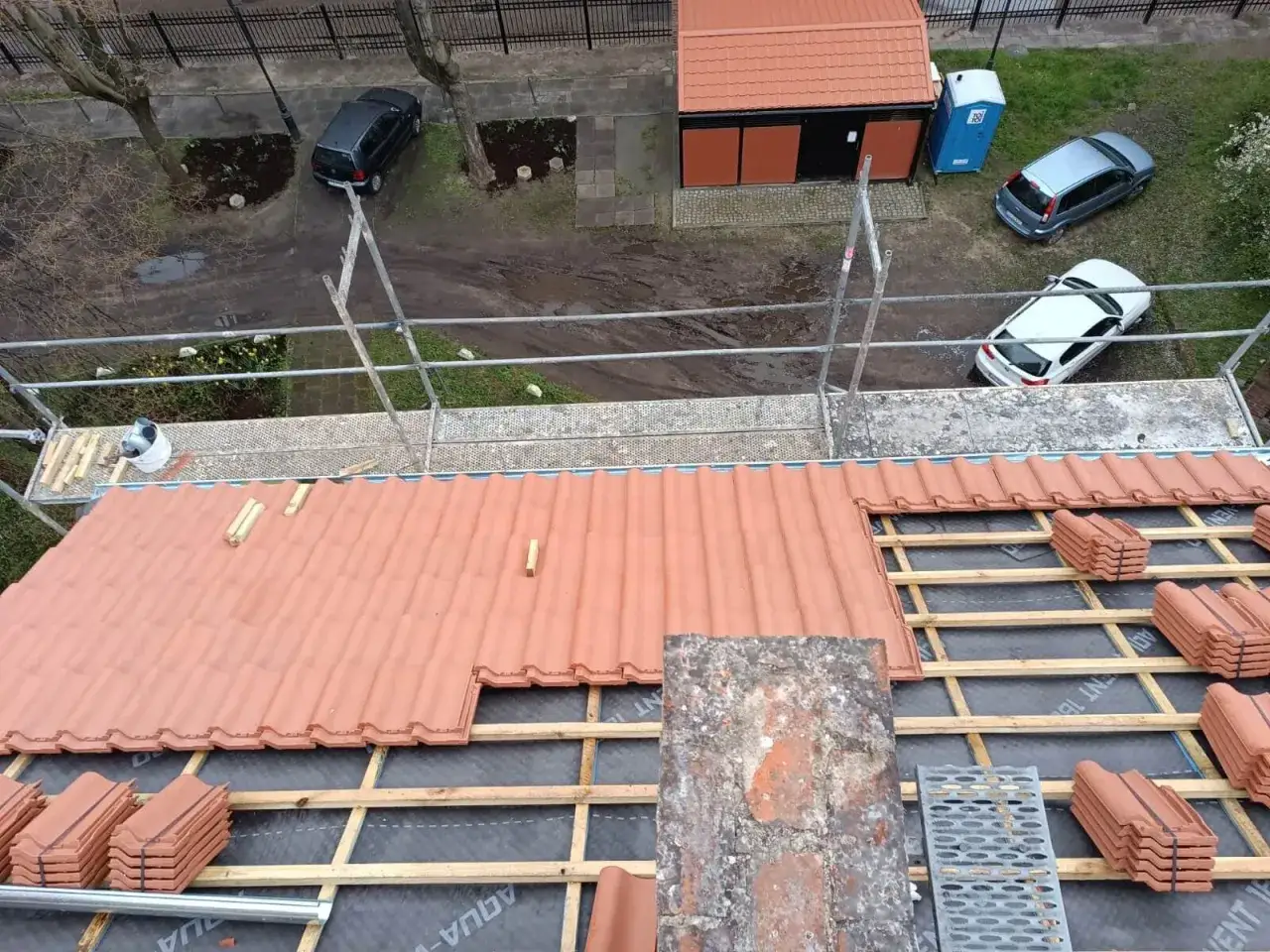 Trwa wymiana dachu. Nowe dachówki czekają na ułożenie na rusztowaniach. W tle widać samochody i budynek.